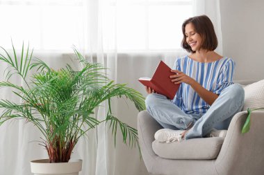 Evdeki koltukta kitap okuyan güzel genç bir kadın.