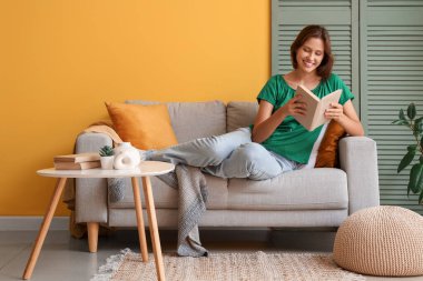 Sarı duvarın yanındaki kanepede kitap okuyan güzel bir kadın.