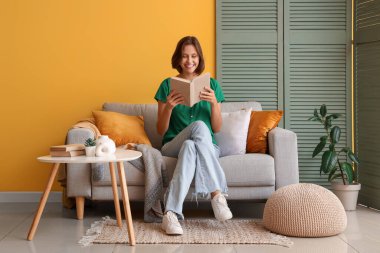 Sarı duvarın yanındaki kanepede kitap okuyan güzel bir kadın.