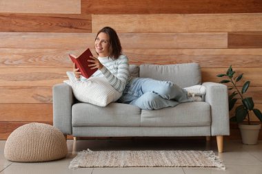 Ahşap duvarın yanındaki kanepede kitap okuyan güzel bir kadın.