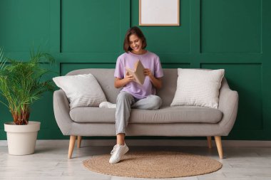 Yeşil duvarın yanındaki kanepede kitap okuyan güzel bir kadın.