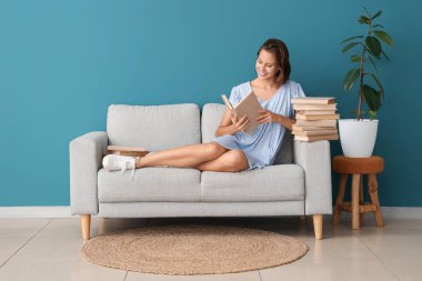 Mavi duvarın yanındaki kanepede kitap okuyan güzel bir kadın.