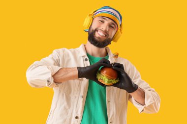 Siyah eldivenli genç adam sarı arka planda lezzetli bir burger tutuyor.