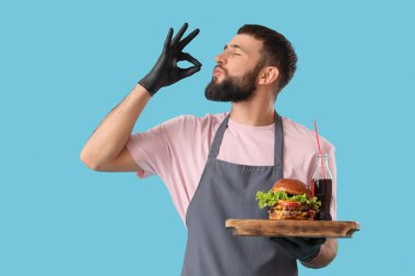 Genç aşçı, ahşap tepside lezzetli bir burger ve mavi arka planda kola tutuyor.