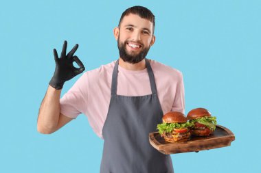 Genç aşçı ahşap tepsiyi lezzetli hamburgerlerle tutuyor ve mavi arka planda iyi görünüyor.