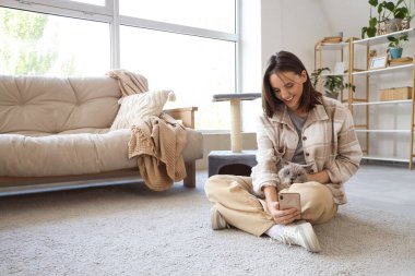 Şirin İngiliz Shorthair kedisi olan genç bir kadın evdeki halıda cep telefonu kullanıyor.