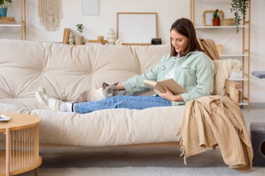 Şirin İngiliz Shorthair kedisi olan genç bir kadın evdeki kanepede kitap okuyor.