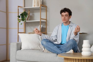 Handsome young man meditating on sofa at home