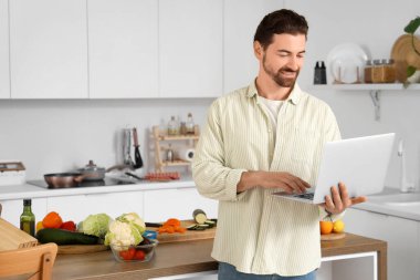 Mutfakta dizüstü bilgisayarı olan yakışıklı sakallı adam.