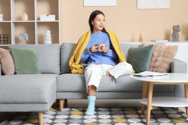 Açık mavi örgü süveter ve battaniyeli ve evdeki kanepede oturan bir fincan çayla güzel genç bir kadın.