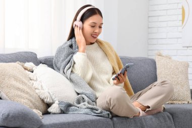 Beyaz örgü süveter, battaniye, kulaklık ve cep telefonlu güzel bir kadın evdeki koltukta oturuyor.