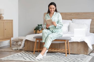 Evde yeşil pijamalı genç bir kadın kitap okuyor.