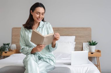 Evde yeşil pijamalı genç bir kadın kitap okuyor.