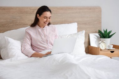 Çizgili pijamalı genç bir kadın evde dizüstü bilgisayar kullanıyor.