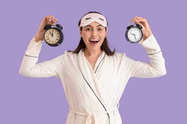 Uyku maskesi ve eflatun arka planda çalar saati olan pijamalı genç bir kadın.