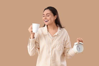 Üzerinde kahve fincanı ve bej arka planda çalar saati olan çizgili pijamalı genç bir kadın.