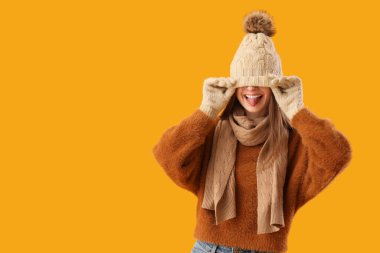 Sarı arka planda dilini gösteren şık, sıcak elbiseli güzel genç bir kadın.