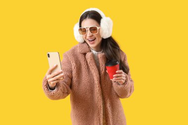 Sarı arka planda kahve fincanıyla selfie çeken, kışlık elbiseli şık genç bir kadın.