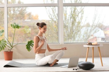 Bilgisayarlı genç bir kadın pencerenin yanında meditasyon yapıyor.