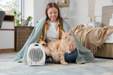 Ekoseli genç kadın ve Maine Rakun kedisi yatak odasındaki elektrikli fan ısıtıcısının yanında oturuyor.