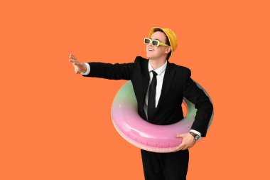 Güneş gözlüklü ve şişme yüzüklü bir ofis çalışanı turuncu arka planda tatil hayali kuruyor.