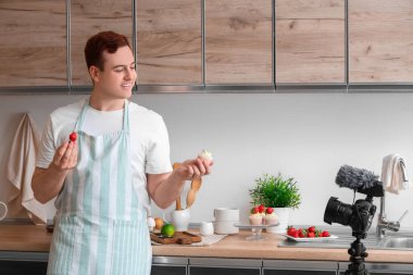 Tatlı kekleri ve taze çilekleri olan genç bir adam mutfakta yemek videosu kaydediyor.