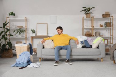 Stresli genç adam evde dağınık giysilerle kanepede oturuyor.