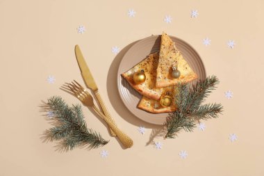 Tabakta lezzetli pizza dilimleri, çatal bıçak takımı, köknar dalları ve bej arka planda Noel topları olan masa seti.