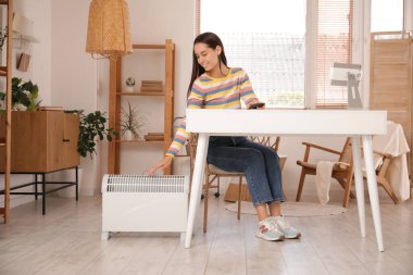 Oturma odasındaki elektrikli ısıtıcının yanındaki masada oturan genç kadın.