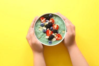 Bir kase lezzetli spirulina smoothie ve sarı arka planda böğürtlenli kadın elleri.