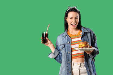 Lezzetli burger tabağı ve yeşil arka planda bir bardak kola tutan güzel bir kadın.