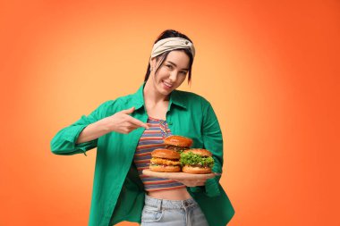 Güzel genç bir kadın turuncu arka planda lezzetli burgerlerle ahşap tahtayı işaret ediyor.
