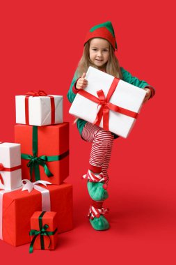 Kırmızı arka planda kutuları olan Noel cücesi kostümlü şirin bir kız.