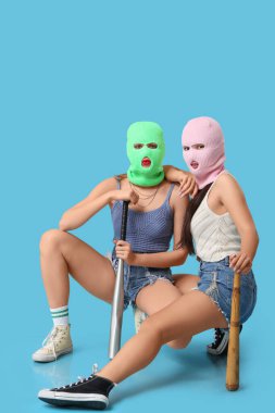 Beautiful young stylish women in balaclavas with baseball bats sitting on blue background clipart