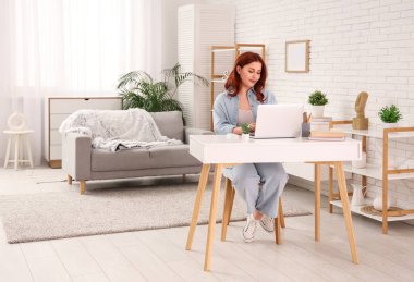 Evdeki masada dizüstü bilgisayar kullanan genç bir kadın.