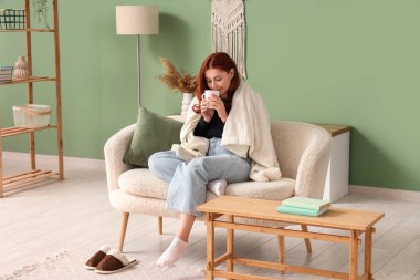 Sıcak ekoseli genç kadın evdeki kanepede çay içiyor.