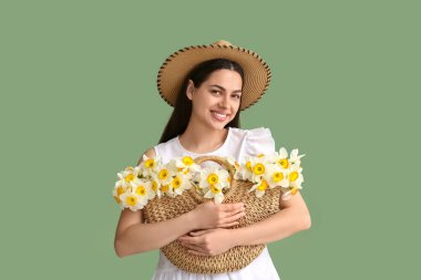 Yeşil arka planda nergis çiçekleri olan hasır bir çanta tutan güzel bir genç kadın.