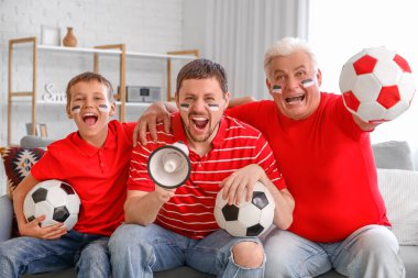 Babası ve oğluyla birlikte oturma odasında megafon ve futbol toplarıyla futbol takımı için tezahürat yapan bir adam.