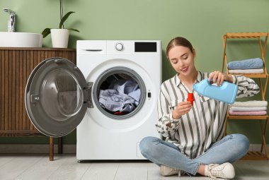 Çamaşır odasında çamaşır makinesinin yanında deterjanı olan genç bir kadın.