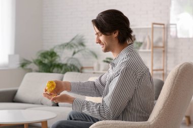 Handsome young happy man with jar of fish oil capsules sitting on sofa at home