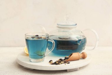 Çaydanlıklı beyaz ahşap tepsi, bir fincan mavi organik çay ve hafif arkaplanda kepçeyle bezelye çiçeği çayı.