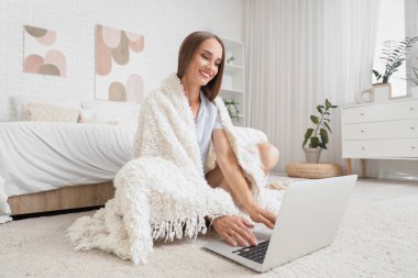 Ekoseli genç bir kadın yatak odasında dizüstü bilgisayar kullanıyor.