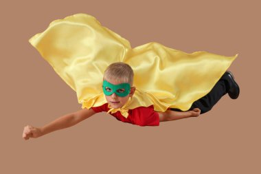 Kahverengi arka planda uçan süper kahraman gibi giyinmiş mutlu okul çocuğu.