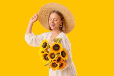 Hasır şapkalı, şık makyajlı ve sarı arka planda bir buket ayçiçeği olan genç ve güzel bir kadın.
