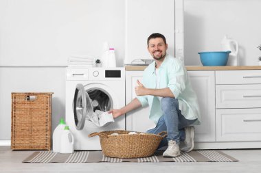 Genç adam çamaşır makinesinden temiz çamaşır alıyor ve evde baş parmağını kaldırıyor.