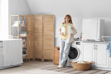 Çamaşır makinesinin yanında çamaşır deterjanı şişeleri olan genç bir kadın.