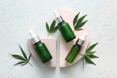 Kenevir kozmetik yağı şişeleriyle dekoratif podyumlar ve açık mavi arka planda yapraklar