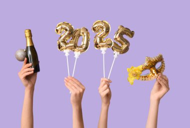 Folyo balonlardan, bir şişe şampanyadan, Noel balosundan ve eflatun arkaplanlı karnaval maskesinden yapılmış 2025 figürlü kadın elleri.