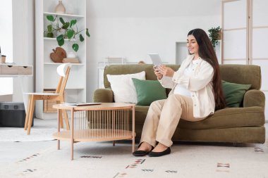 Hamile iş kadını evdeki kanepede tablet bilgisayarla çalışıyor.