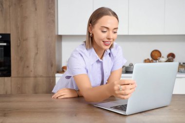 Dizüstü bilgisayarlı genç bir kadın mutfaktaki masada sohbet ediyor.